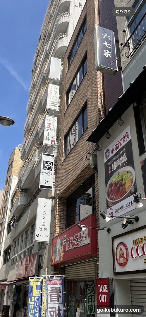 Vertical view of multiple shop fronts on a city street under clear blue sky. Visible signage includes '六七八家', 'WAVEZ 2F', 'KLINK', and various other banners. One prominent poster advertises food items with Japanese text. Store entrances are partially visible at ground level.