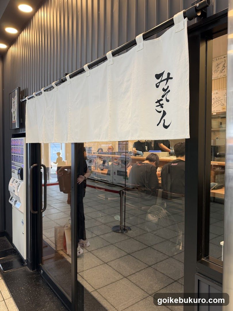 The image shows the exterior entrance of a restaurant, featuring a white noren (door curtain) hanging above the glass doors. The noren has Japanese characters written on it. Inside, customers are seated at tables, and staff members can be seen working behind the counter. A vending machine is visible to the left of the entrance.