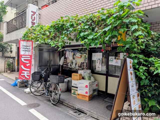 A small storefront entrance covered by lush green vines, featuring a vertical red sign with Japanese characters, stacked cardboard boxes near the door, bicycles parked outside, and an A-frame menu board leaning against the wall.