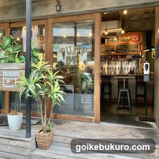 Wooden storefront with glass doors and windows displaying 'PUMP ART BAR' signage. Interior shows beer taps, bar stools, and hanging lights. Plants in pots on wooden deck outside.