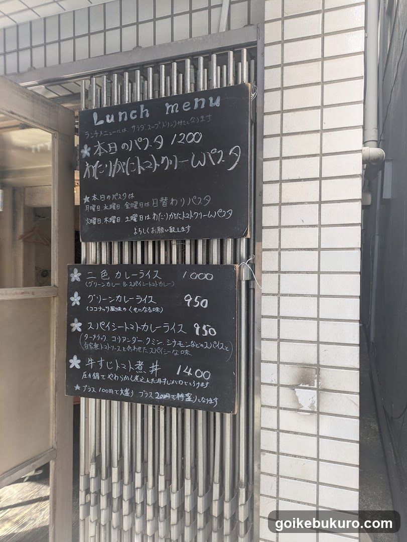 A photo of a shop's exterior entrance with two black chalkboard signs mounted on a metal gate. The top sign displays 'Lunch menu' and lists items like 'Today's Pasta 1200'. The bottom sign lists rice dishes such as 'Two-color Curry Rice 1000', 'Green Curry Rice 950', 'Spicy Tomato Curry Rice 950', and 'Udon 1400'. The background shows white tiled walls and part of the building facade.