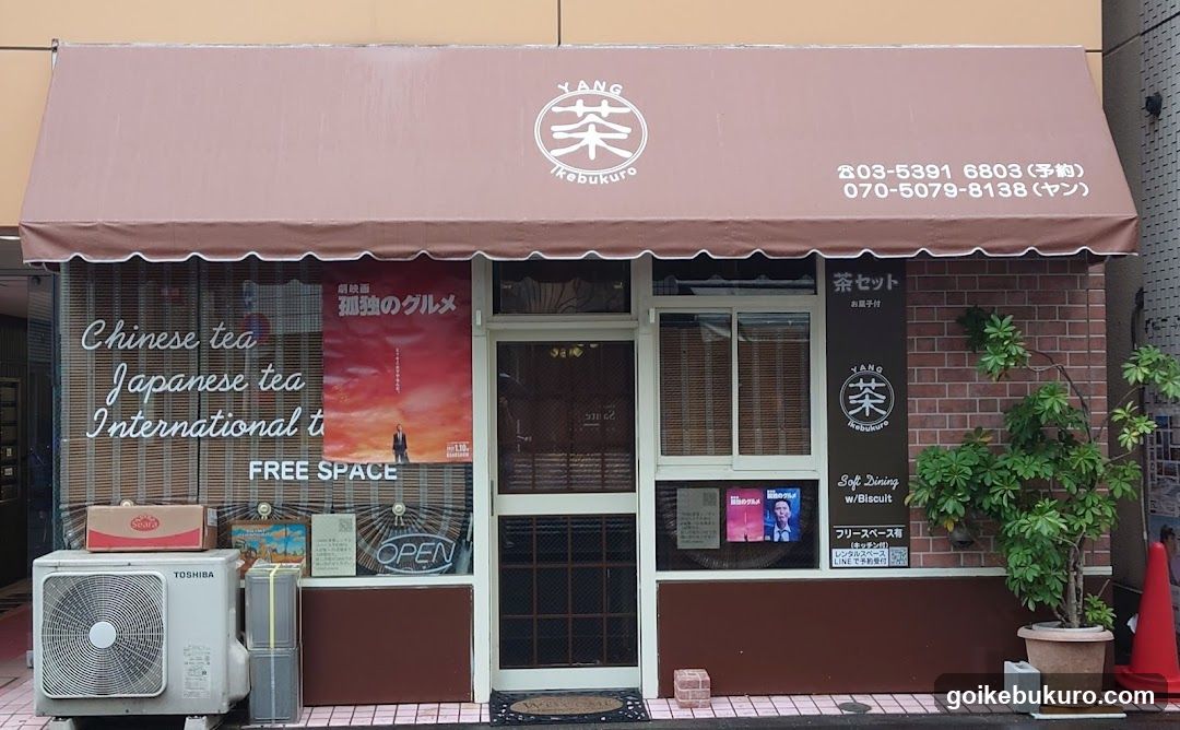 A storefront with a brown awning displaying the logo 'YANG' and Japanese characters. The building features large windows with posters advertising Chinese tea, Japanese tea, and international tea. A sign lists phone numbers and mentions tea sets and biscuits. An air conditioner unit is visible on the left side of the entrance.