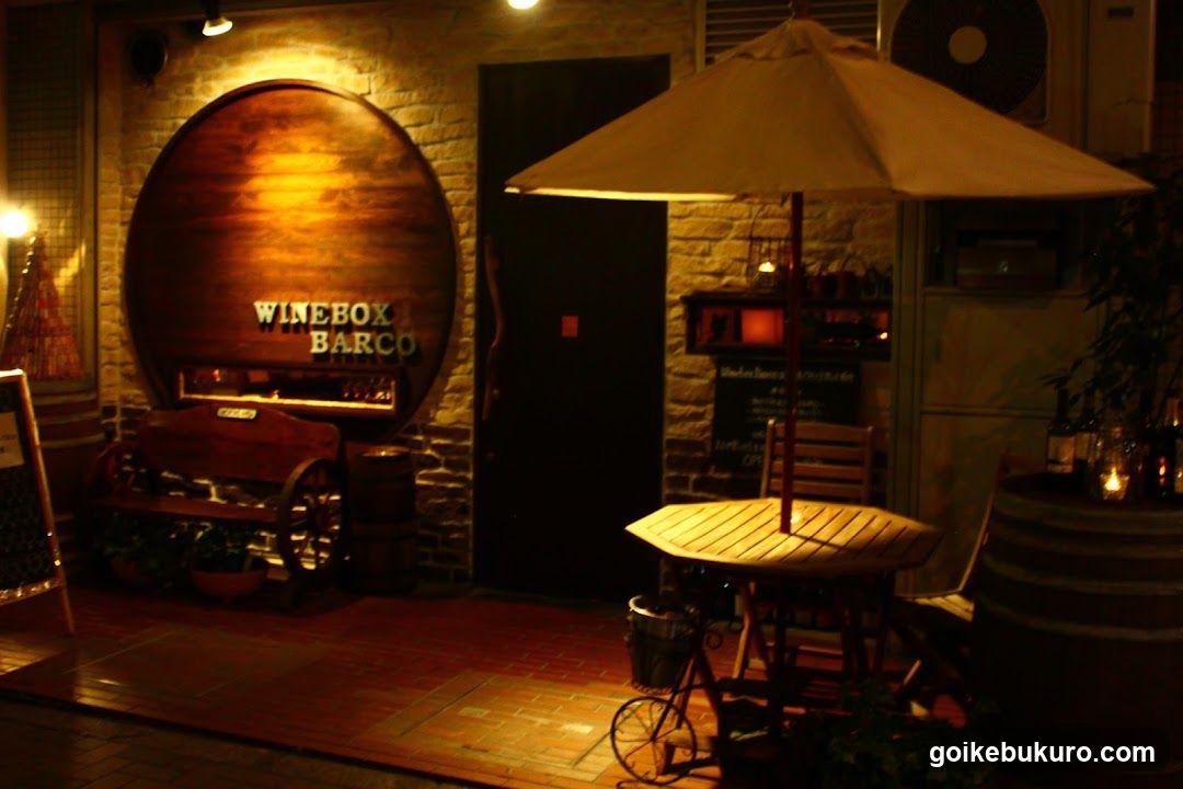 A warmly lit storefront with a large circular wooden sign reading 'WINEROOM BARCO' mounted on a brick wall. A small outdoor seating area features a table under an umbrella, flanked by barrels and potted plants. The entrance is dark, suggesting evening ambiance.