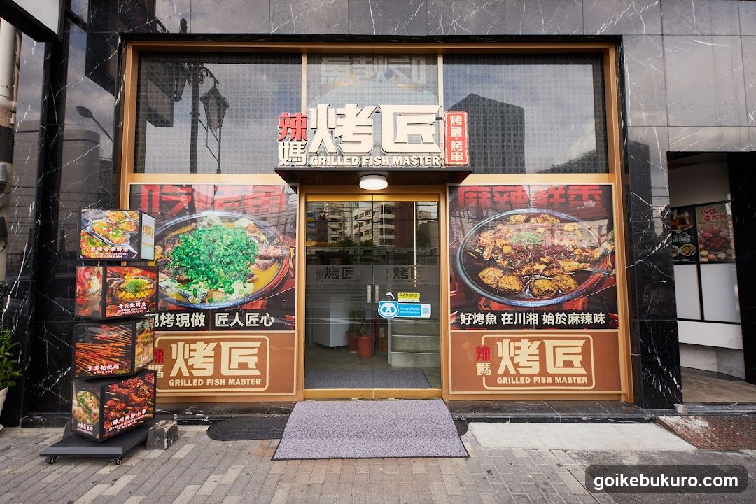 A modern shop front with large glass windows and doors, featuring prominent signage reading 'GRILLED FISH MASTER' in both Chinese and English. The facade includes decorative posters showcasing spicy fish dishes, with one poster highlighting a green-topped dish and another displaying a red-spiced fish meal. A vertical display stand to the left holds packaged food items. The entrance has a gray mat on the sidewalk.