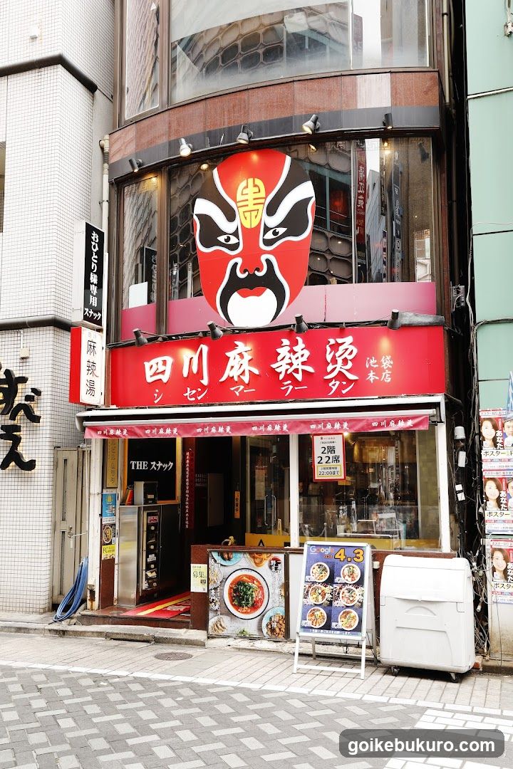 A street-level view of a Sichuan Malatang restaurant with a large red signboard featuring Chinese characters and Japanese katakana. A prominent red mask logo is displayed above the entrance. The storefront includes glass windows, an open doorway, and various signs advertising menu items like '2-plate' and '3-plate'. A white washing machine sits outside near the sidewalk.