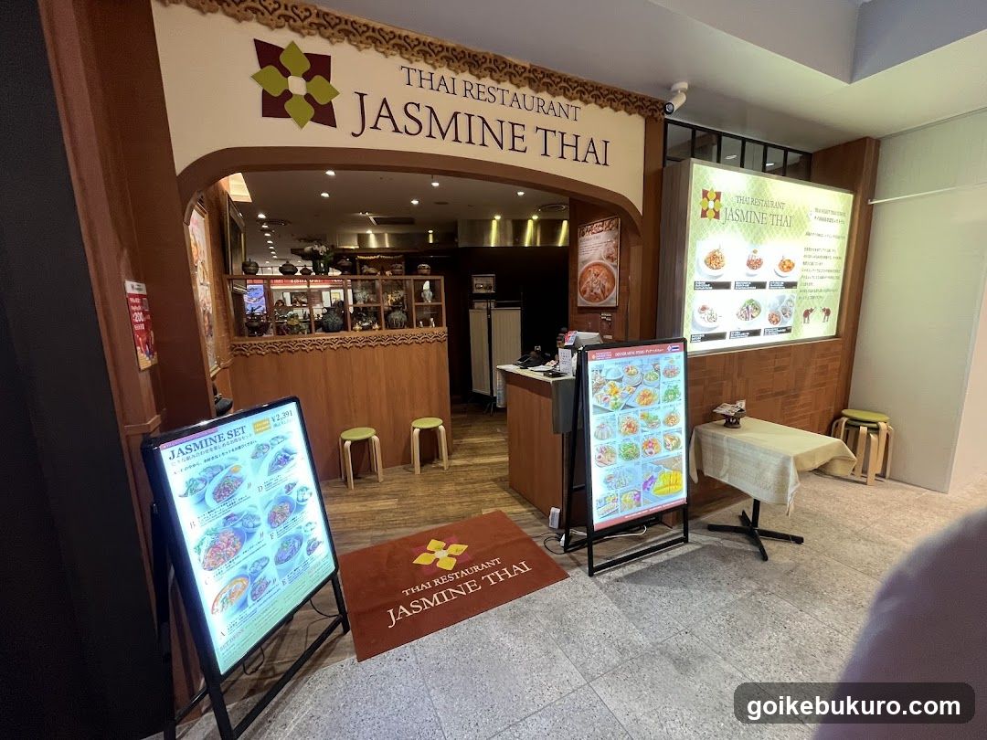 Entrance to a Thai restaurant with signage reading 'THAI RESTAURANT JASMINE THAI' above the archway. A digital menu board stands outside, displaying various dishes and prices. Inside, wooden decor and shelves are visible. Additional promotional posters and a small table with stools are present near the entrance.