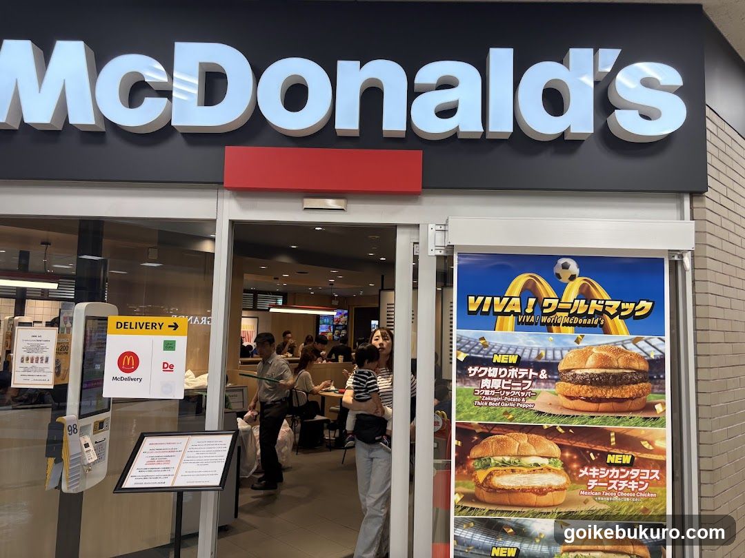 Photo of the physical exterior entrance of a McDonald's restaurant, showing large white signage on a black background above glass doors. A red rectangular sign is mounted below the main logo. Inside, customers are seated at tables and standing near counters. On the right side, there is a vertical poster advertising new menu items in Japanese, including 'VIVA! ワールドマック' with images of burgers like 'Sakura Potato & Beef Patty Burger' and 'Mexican Chicken Cheeseburger'. To the left, a self-service kiosk displays delivery options for McDonald’s and De’Longhi coffee.