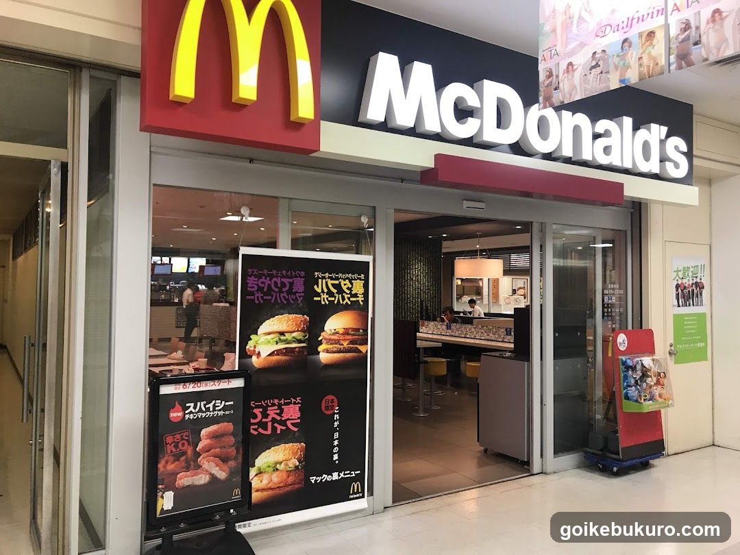 A photo of a McDonald's storefront with visible signage, including the iconic golden arches logo and 'McDonald's' lettering. The entrance features glass doors leading into the dining area. Promotional posters display menu items like burgers and chicken sandwiches. A red promotional stand is positioned near the entrance.