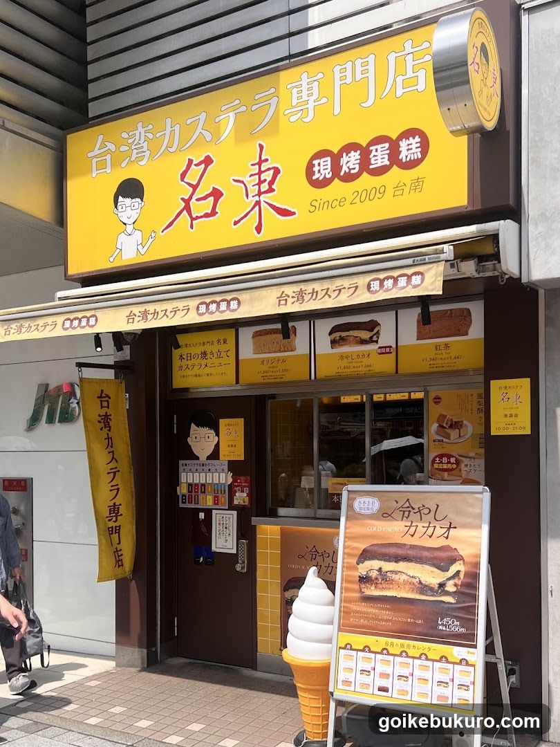 A storefront with a yellow signboard displaying '台湾カステラ専門店' and '名東'. The shop features a menu board outside, showcasing various castella cake products like strawberry, chocolate, and red bean flavors. There is also an ice cream cone display in front of the entrance.