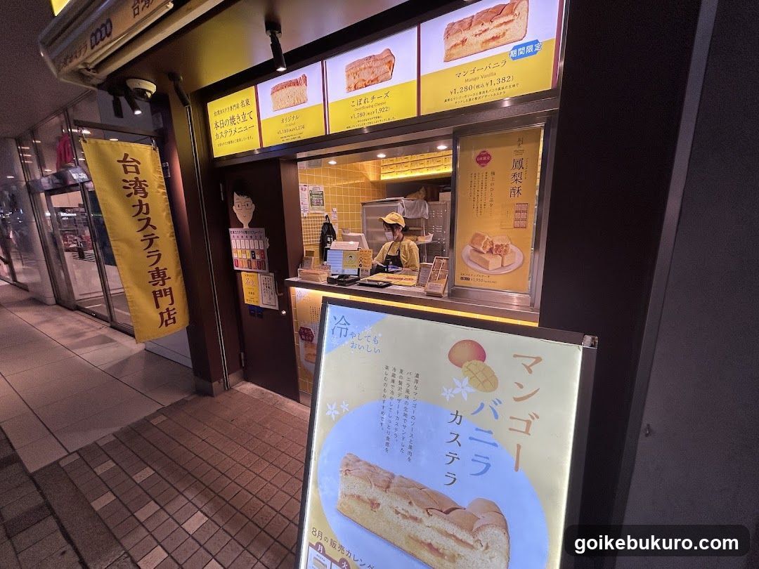 Nighttime exterior of a shop with yellow signage and illuminated menu boards displaying castella cake products. A staff member in uniform is visible inside the counter area. Vertical banners and horizontal signs feature Japanese text promoting Taiwan-style castella.