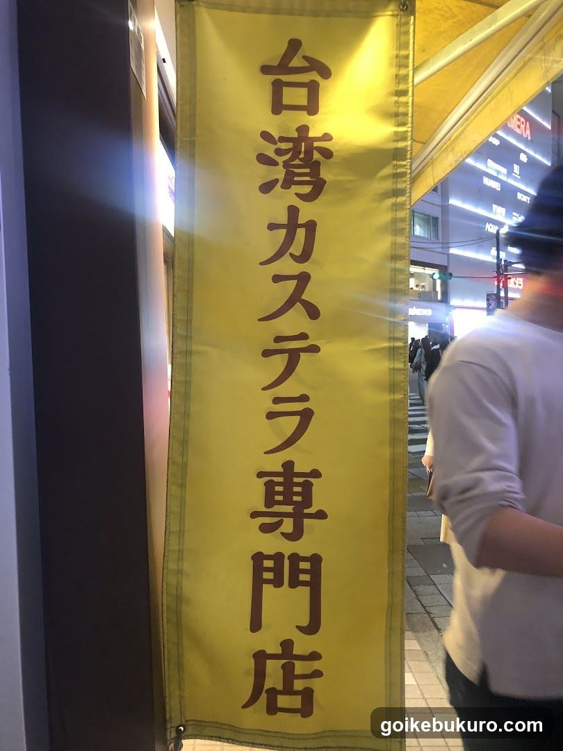 Vertical yellow banner with brown Japanese and Chinese characters reading '台湾カステラ専門店' (Taiwan Castella Specialty Store), hanging at the entrance of a shop in an urban street setting.