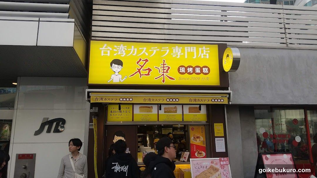 A storefront with a bright yellow sign displaying Japanese and Chinese characters, featuring an illustration of a person and the name 'Meido' (名東). The shop sells castella cake, as indicated by the signage. There are people standing in front of the store.