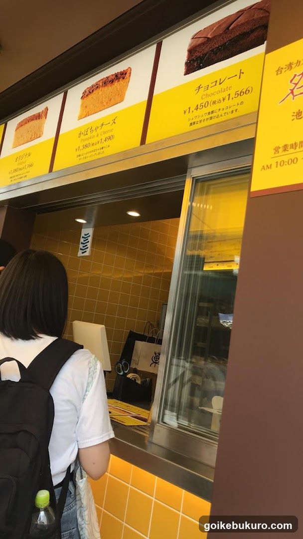 A person stands at a cafe counter, facing the service window. Above the window is a menu board displaying three cake items with prices in yen. The interior behind the counter has yellow tiled walls and glass display cases.