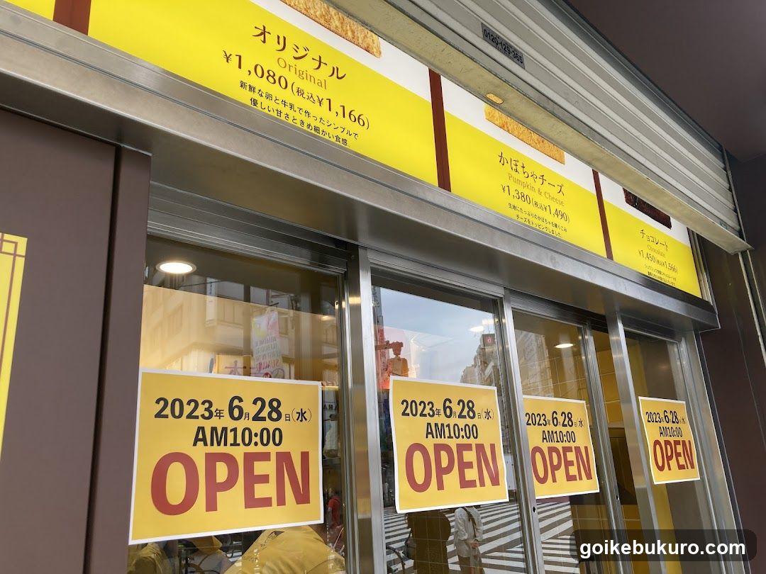The image shows the exterior of a shop entrance with glass doors. Yellow signs above the door display prices in Japanese Yen, including 'Original' at ¥1,080 (tax included ¥1,166) and 'Kobachi Cheese' at ¥1,380 (tax included ¥1,490). Below, yellow posters indicate opening hours: June 28, 2023, from AM10:00 to AM10:00.