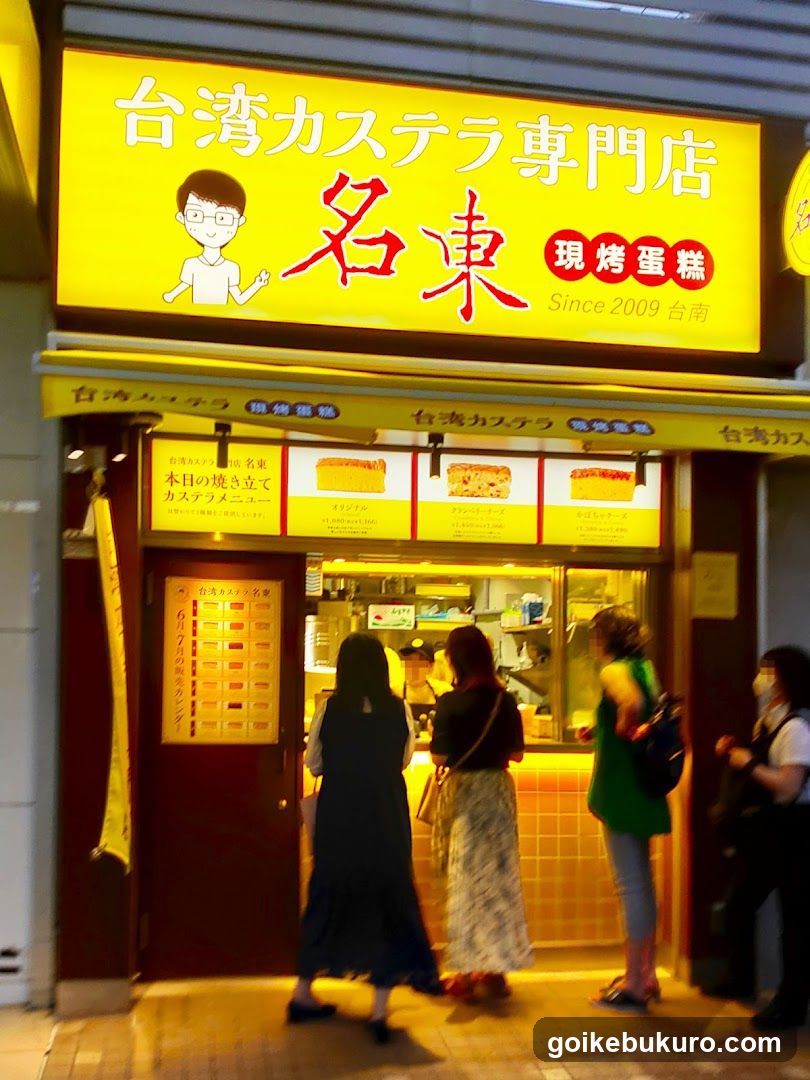 A brightly lit storefront with a large yellow sign reading '台湾カステラ専門店 名東' (Taiwan Castella Specialty Store Meido) and '現烤蛋糕 Since 2009'. Below, three customers stand at the counter ordering from behind glass. A vertical menu board is visible on the left door.