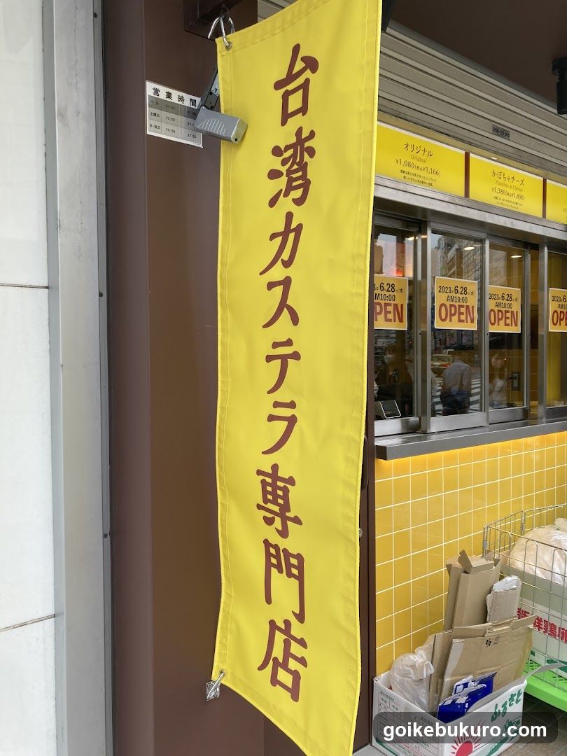 A yellow vertical banner with Japanese and Chinese characters hangs on the storefront, indicating a specialized shop for Taiwan-style castella cake. The entrance features glass doors with 'OPEN' signs and dates displayed above them. Inside, yellow tiled walls are visible along with stacked cardboard boxes near the entrance.