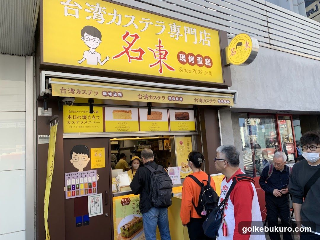 A storefront with a yellow sign displaying '台湾カステラ専門店 名東' and '現烤蛋糕 Since 2009'. A circular logo is mounted on the wall. Below, there are menu boards showing castella cake products. People are standing in line at the entrance.