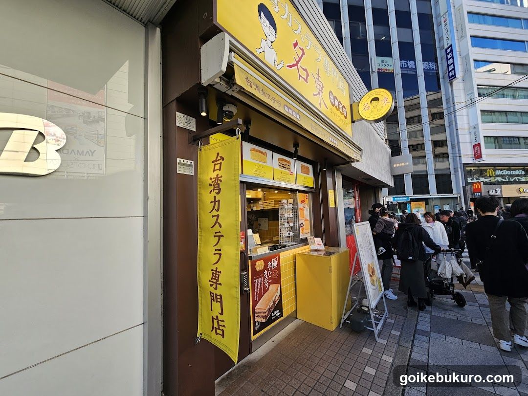 A street-level view of a shop front with yellow signage and banners advertising Taiwan-style castella cake. A vertical banner reads '台湾カステラ専門店' (Taiwan Castella Specialty Shop). The storefront features glass windows displaying baked goods, including layered cakes and sandwiches. People are walking by on the sidewalk in an urban setting.