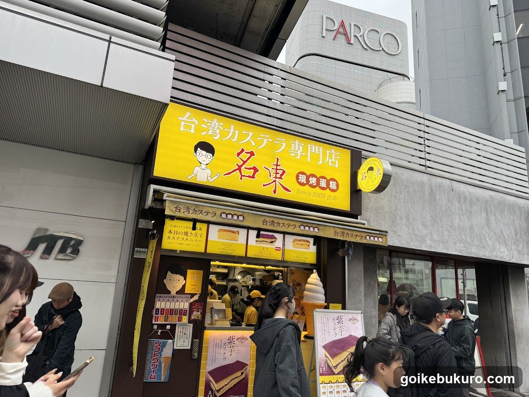 A street-level view of a shop front with a bright yellow sign reading '台湾カステラ専門店 名東' (Taiwan Castella Specialty Store Meido). The storefront features glass windows displaying baked goods, including layered cakes and cream puffs. A queue of customers is visible outside, waiting to enter or purchase items. Posters advertise purple-layered castella cakes. The building facade includes modern architectural elements like horizontal slats and a large PARCO sign in the background.