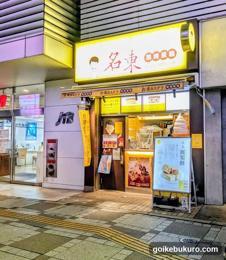 A brightly lit storefront with a yellow sign displaying '名東 環烤蛋糕' and 'Since 2009'. The entrance features glass doors, a menu board outside showing cake slices, and vertical banners hanging on the sides. Adjacent is another shop with signage.