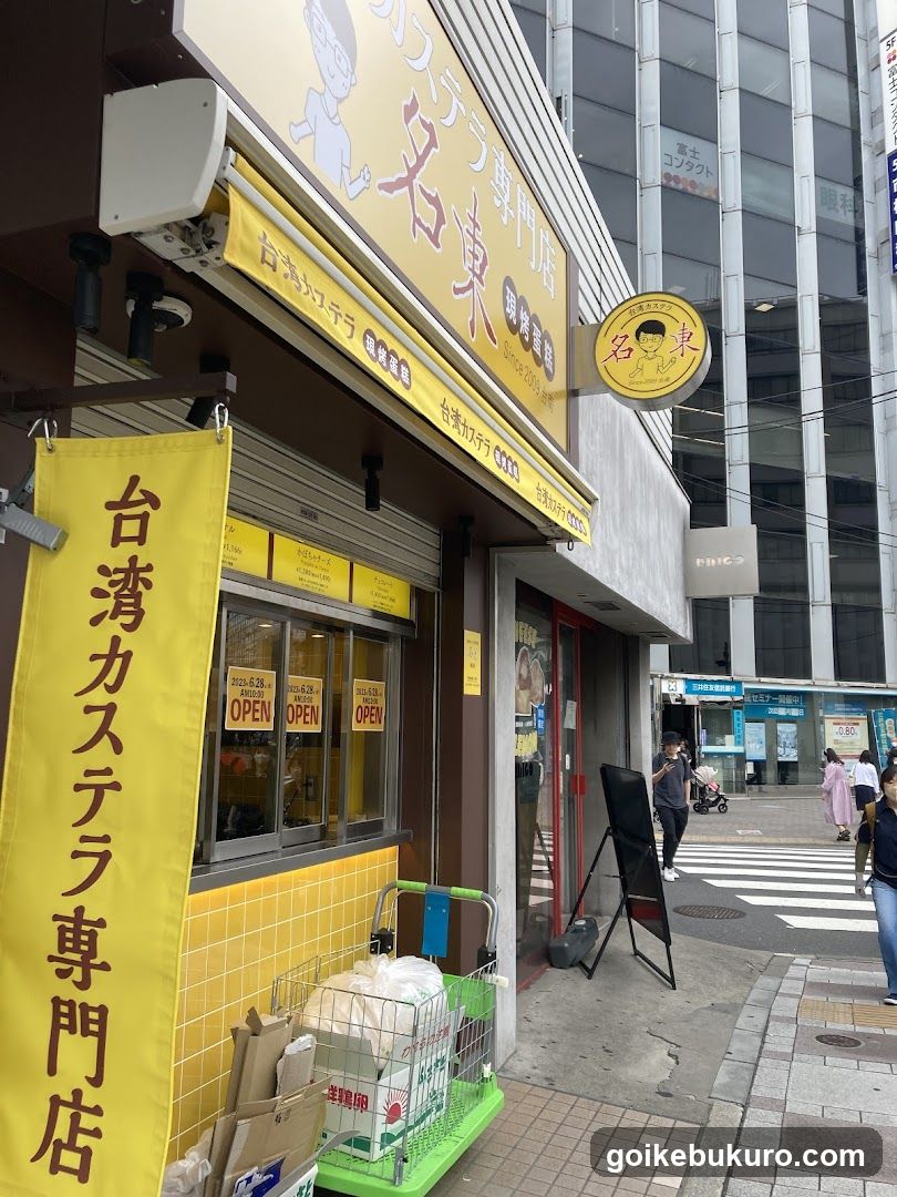 A street-level view of a shop with yellow signage reading '台湾カステラ専門店' (Taiwan Castera Specialty Store). A vertical banner hangs on the left, and a circular sign is mounted above the entrance. The storefront features glass windows displaying 'OPEN' signs inside. A green shopping cart filled with white bags sits near the entrance. Pedestrians walk by in the background.