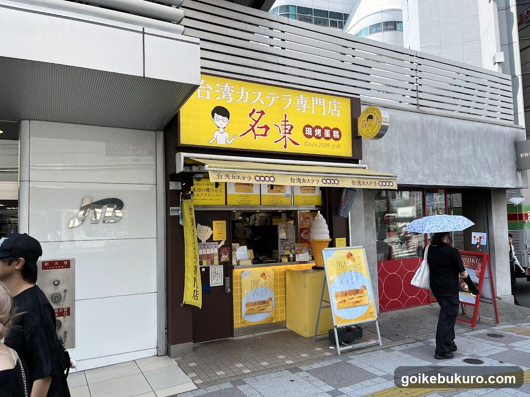 A street-level view of a shop with a yellow sign reading '台湾カステラ専門店' (Taiwan Castella Specialty Store). The storefront features a counter with a display of castella cake and other baked goods. A person holding an umbrella stands nearby, and another individual is partially visible on the left side.