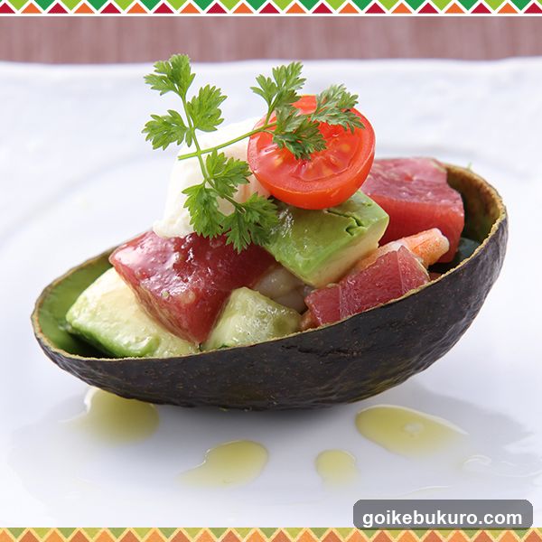 A beautifully presented dish featuring diced raw tuna and avocado cubes arranged in a hollowed-out avocado shell, garnished with fresh parsley and a halved cherry tomato. The salad is drizzled with olive oil on a white plate.