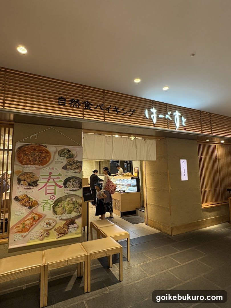 The image shows the entrance of a shop with wooden slat walls and signage reading '自然食バイキング' (Natural Food Buffet) in Japanese characters above the door. A large poster on the left displays various food items under the heading '春の香' (Scent of Spring), featuring dishes like rice bowls, salads, and breads. Inside, customers are seen near a serving counter where staff members work behind glass display cases. Wooden benches line the walkway outside.