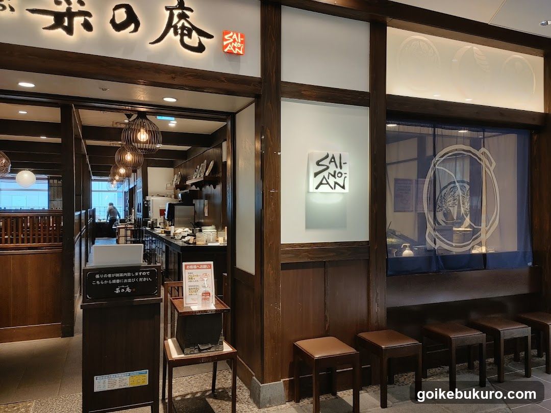 A traditional Japanese shop front with wooden beams and a blue curtain featuring a circular design. The entrance is visible, leading to an interior space with hanging lights and kitchen equipment. A sign on the wall reads 'SAI NO MON', and there are small stools outside.