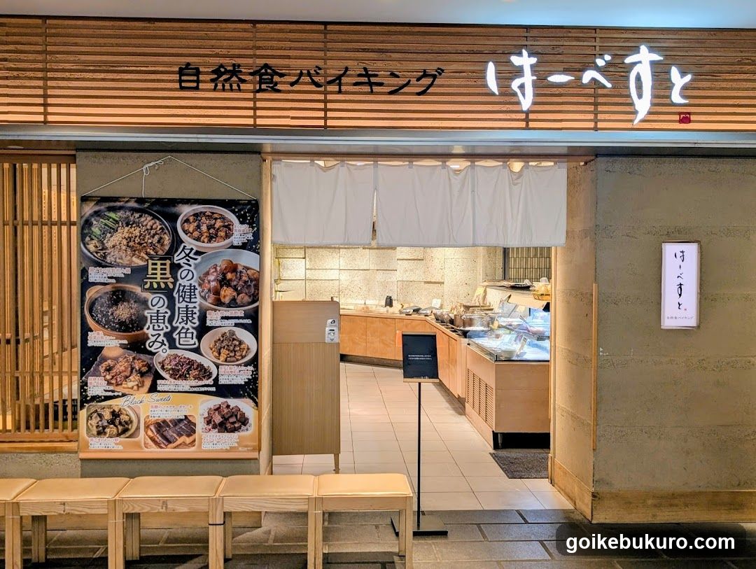 The image shows the exterior entrance of a shop with wooden slat signage displaying Japanese characters and English text 'Natural Food Baking'. A large poster on the left showcases various food items, including dishes like rice bowls and stews, with text highlighting health benefits. Inside, a counter with food display cases is visible, along with a white curtain at the entrance. Wooden benches are placed outside.