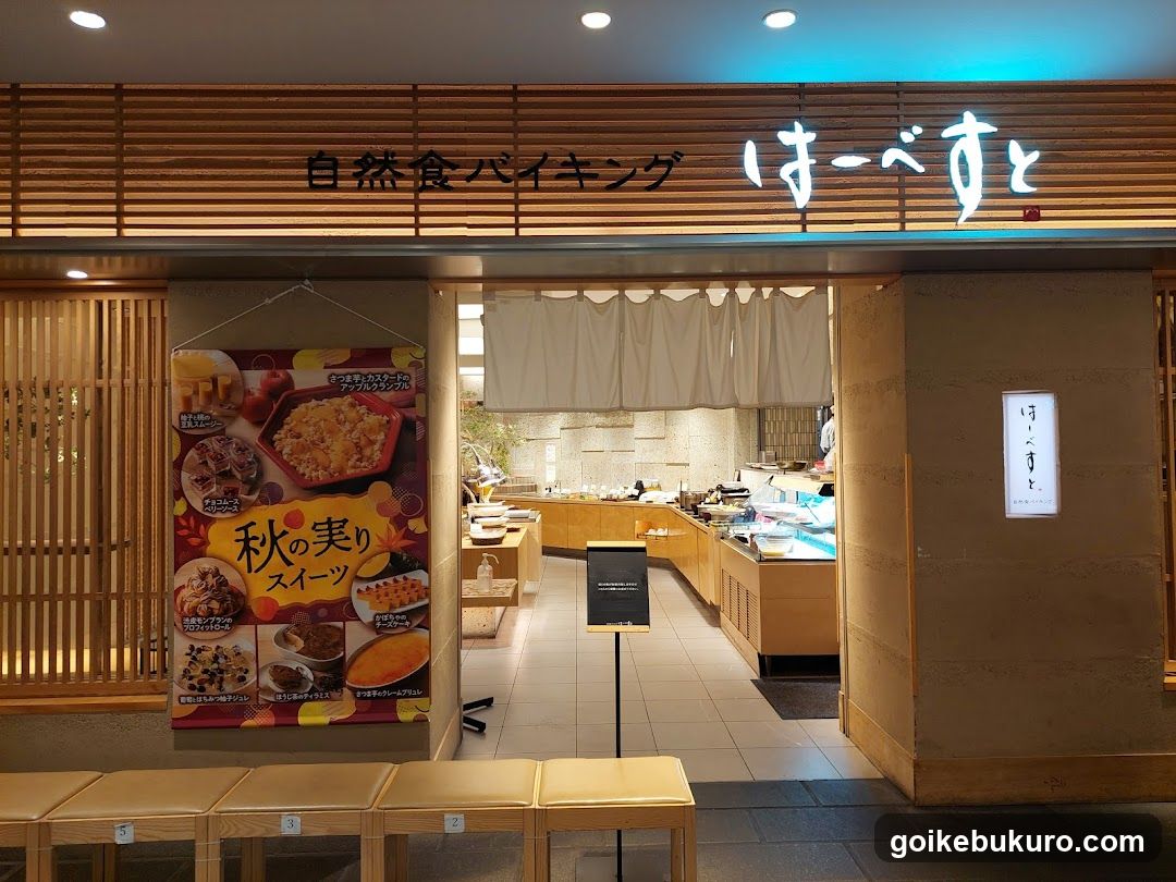 A shop front with wooden slat signage displaying '自然食バイキング' and 'ほーべすと'. A poster advertises autumn sweets, showing various desserts like rice pudding and fruit tarts. The entrance reveals a buffet-style interior with food stations.