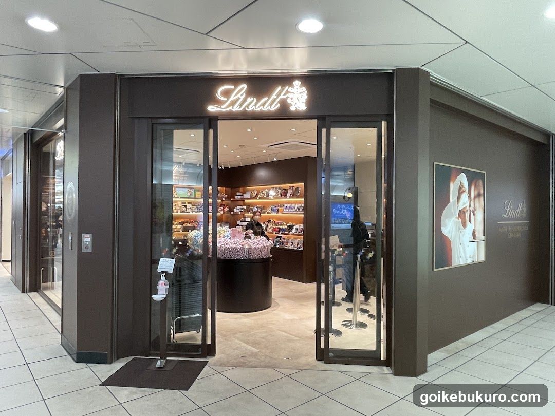 A storefront with a dark brown facade and illuminated 'Lindt' logo above the entrance. Glass doors open to reveal shelves of products inside, including chocolate displays on a central stand.