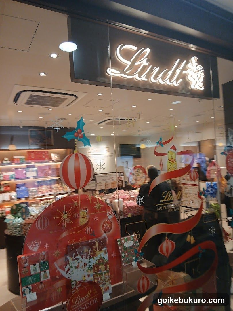 A storefront of a Lindt chocolate shop with illuminated signage above the entrance. The window display features festive decorations including red and white striped ornaments, snowflakes, and holiday-themed posters. Inside, shelves are stocked with various packaged chocolates and confectionery products.