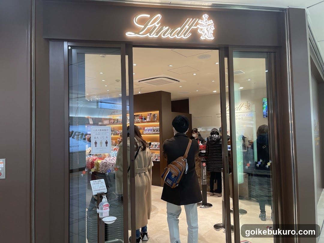 A storefront with illuminated 'Lindt' signage above glass doors. Inside, customers browse shelves of confectionery products near a checkout counter.