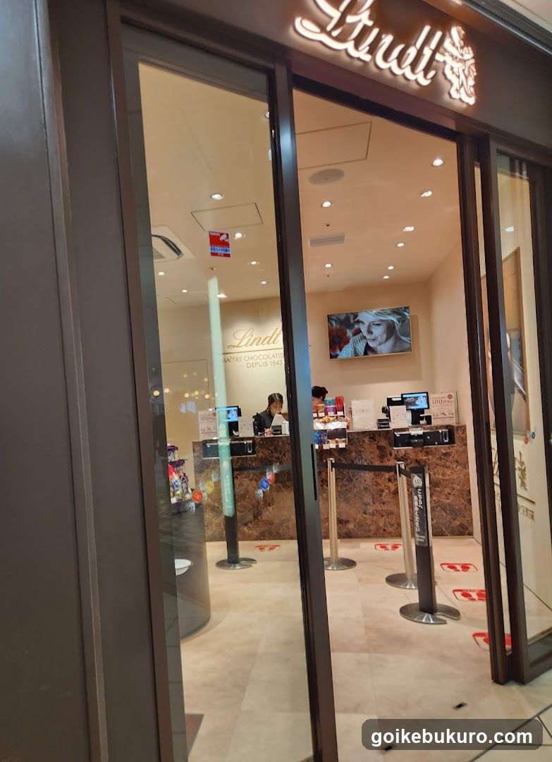 The image shows the exterior entrance of a Lindt chocolate store, with visible signage above the door. Inside, there is a reception desk with staff members attending to customers. A television screen displays an advertisement or promotional content on the wall behind the counter. The interior features warm lighting and marble accents.