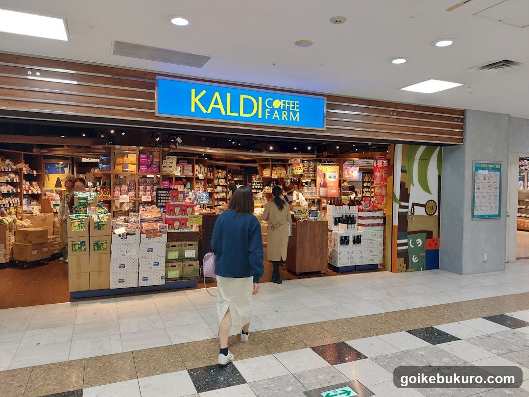 A brightly lit storefront with a blue sign reading 'KALDI COFFEE FARM' above the entrance. The shop displays various products on shelves and stacked boxes near the door. Two people are walking toward the store, one carrying a pink bag. Inside, staff members are visible behind counters.