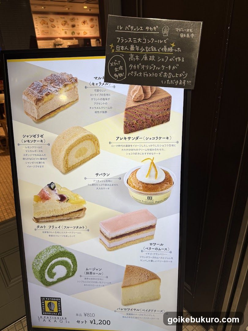 Close-up of a vertical menu board displaying various French-style cakes and pastries with Japanese descriptions, prices, and promotional text on a chalkboard sign above.