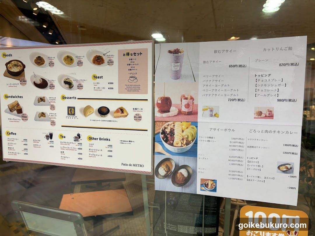 A close-up of a printed menu board displayed on glass, featuring sections for Foods, Toast, Sandwiches, Desserts, Coffee, Tea, and Other Drinks. The right side shows drinks like Berry Asai and Apple Pie, along with a bowl of fruit salad and a curry dish.