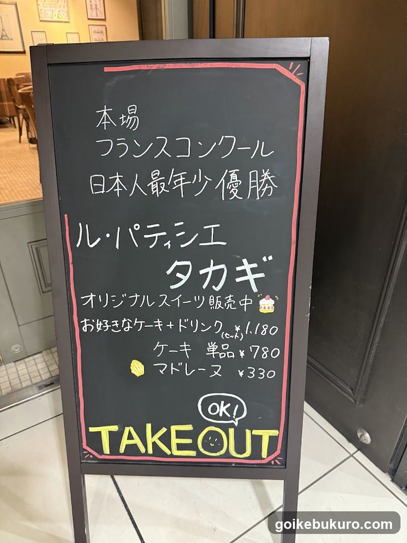 A chalkboard menu board outside a shop advertising French-style cakes and pastries, including Takagi cake and macaron.