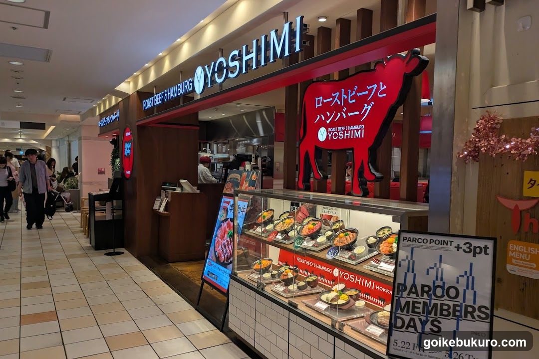 The image shows the exterior of a Yoshimi store, featuring prominent signage with 'ROAST BEEF & HAMBURGER' and Japanese text. A large red cow graphic is visible on the facade. Inside, food items are displayed in glass cases, including various dishes like rice bowls and grilled meats. Staff can be seen behind the counter.