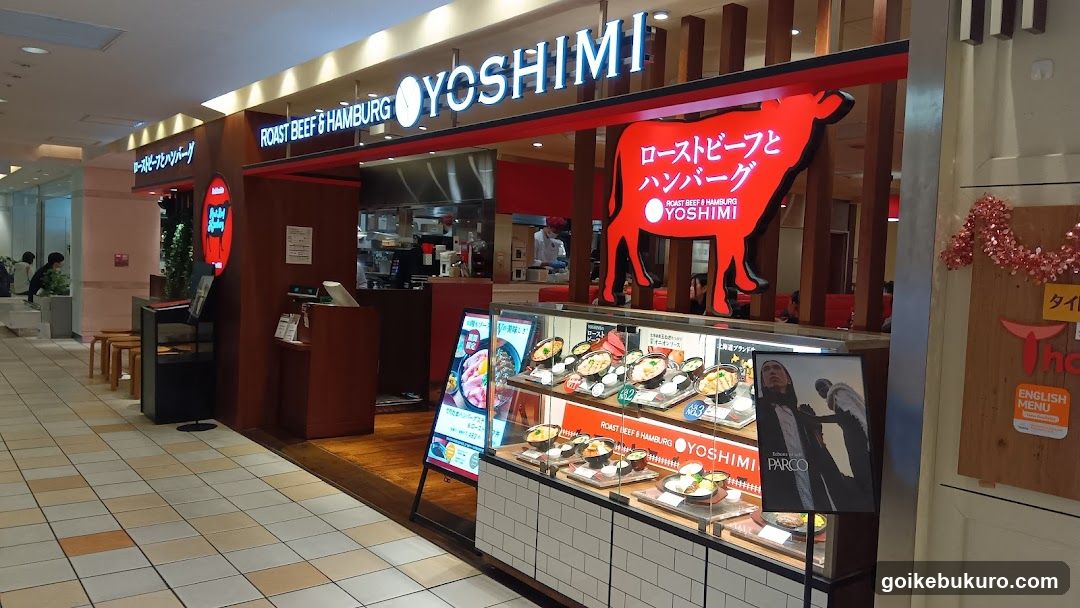 A storefront of Yoshimi, a Japanese restaurant specializing in roast beef and hamburgers. The shop features prominent signage with the name 'YOSHIMI' illuminated above the entrance. A large red silhouette of a cow is displayed on the wall inside the store. Outside, there are promotional posters showcasing food items and a menu board displaying various dishes with prices.