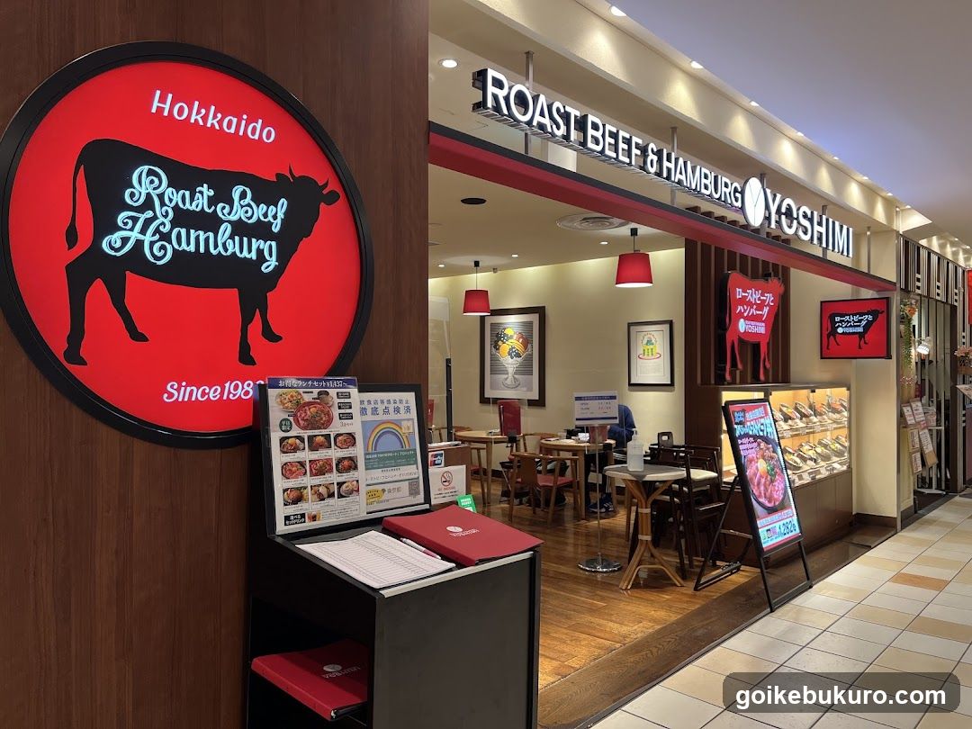 Storefront of a restaurant with signage reading 'ROAST BEEF & HAMBURG YOSHIMI' and a large circular logo featuring a cow silhouette. Interior visible through glass doors, showing tables, chairs, red pendant lights, framed posters on walls, and promotional banners. A menu stand outside displays food items with prices.