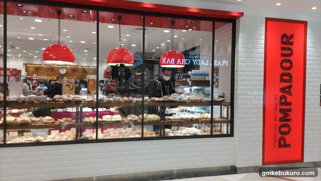 A bakery storefront with a large glass window displaying shelves of bread and pastries. Red pendant lights hang above the display. A vertical red sign on the right reads 'POMPADOUR' in black letters, with smaller text below indicating 'YOKOHAMA MOTOACHI'. Inside, staff are visible behind the counter.