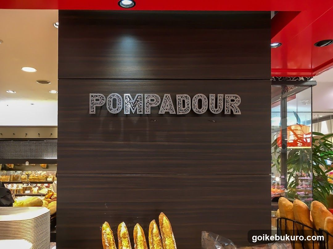 A close-up view of a bakery storefront featuring the word 'POMPADOUR' in stylized, metallic lettering mounted on a dark wood-paneled wall. Below the sign are several baguettes displayed in a wire rack. To the left, shelves with baked goods and bread loaves are visible inside the shop. The ceiling above has recessed lighting and red accents.