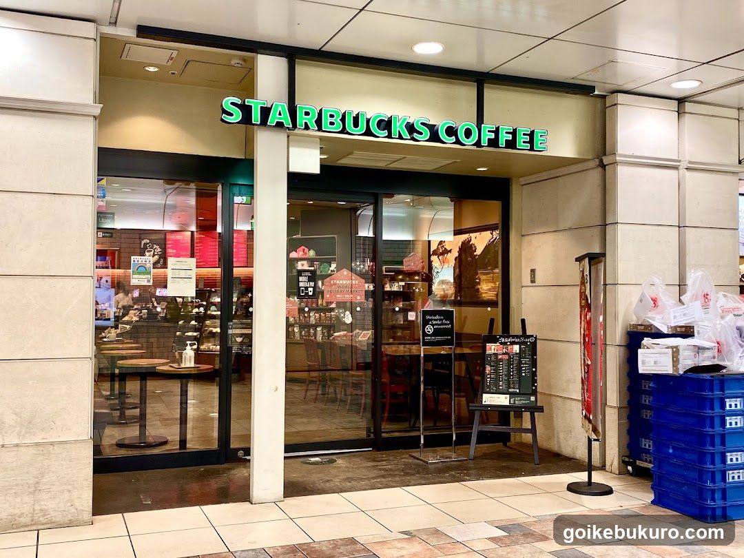 The image shows the exterior of a Starbucks Coffee shop, featuring illuminated green signage above glass doors and windows. Inside, tables and chairs are visible along with shelves stocked with products. Outside to the right, blue crates hold white plastic bags.