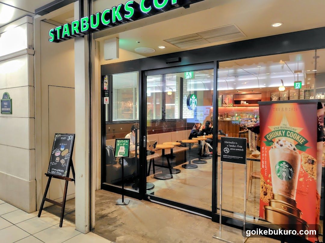 Photo of the physical exterior entrance and storefront facade of a Starbucks coffee shop. The green neon 'STARBUCKS COFFEE' sign is mounted above the glass doors on the building's wall. Inside, customers are seated at tables near the counter. A large poster advertising the 'Chunky Cookie' drink stands to the right of the entrance. A chalkboard menu board with illustrations is placed outside to the left.