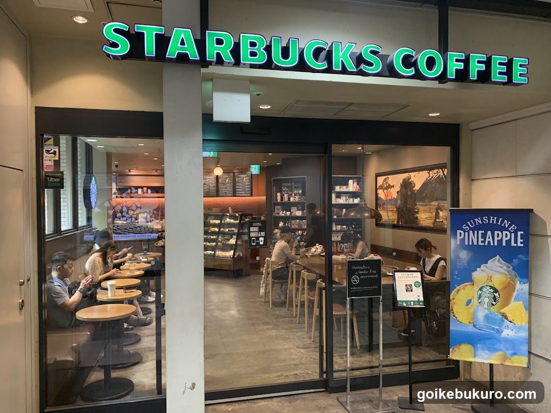 A Starbucks Coffee storefront with a bright green illuminated sign above the entrance. The interior is visible through glass doors, showing customers seated at tables and a display case of baked goods. A promotional poster for 'Sunshine Pineapple' drink stands outside near the entrance.
