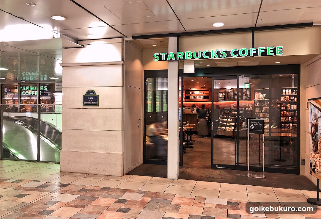 A Starbucks Coffee storefront with illuminated green signage above the entrance. The glass doors and windows reveal the interior, including shelves stocked with products and a counter area. A sign on the wall reads 'PLACE OF INTEREST'. An escalator is visible to the left.