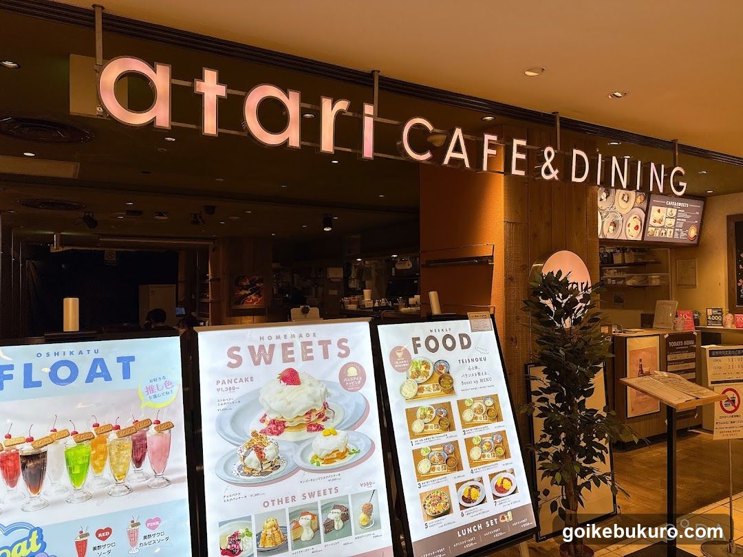 The image shows the storefront of a cafe and dining establishment with illuminated signage reading 'atari CAFE & DINING'. Below, there are three large menu boards displaying various food items including floats, sweets like pancakes and other desserts, and lunch sets.