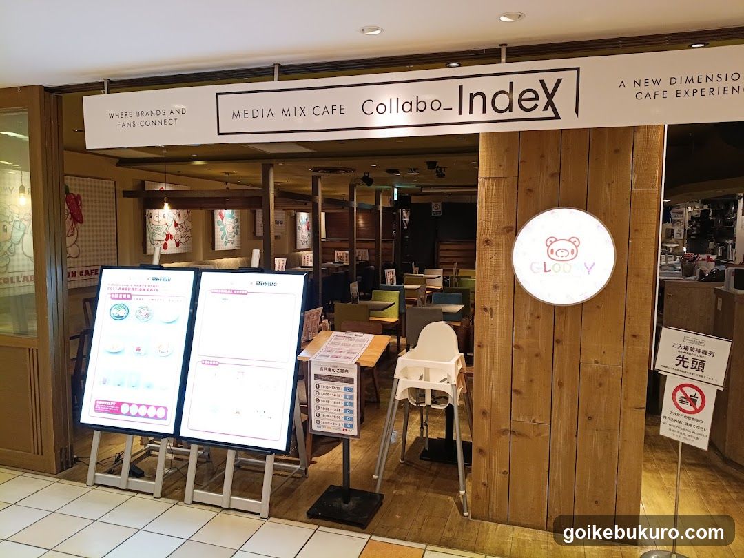 Entrance to Collabo_Index, a media mix cafe featuring wooden paneling and illuminated signage. Visible menu boards display food items with prices. Interior seating area includes high chairs and tables.